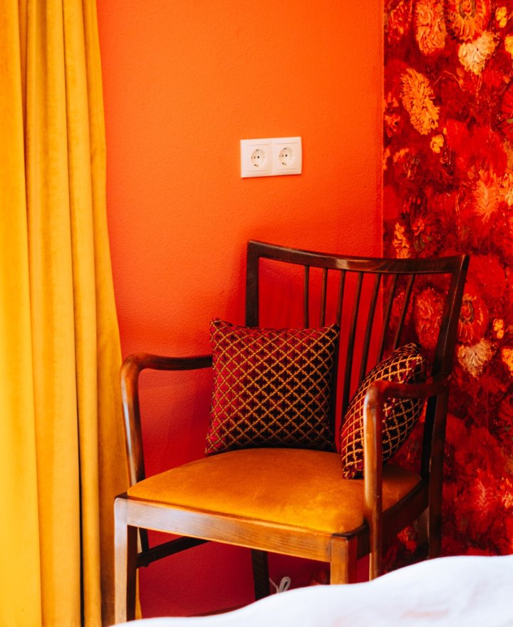 Hotelkamer met feloranje wand, rood bloemenbehang, vintage stoel met okergele zitting en gele gordijnen, door cestsjoukje