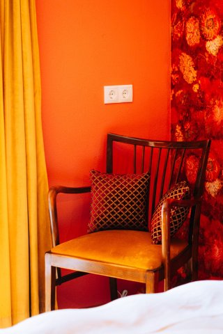 Hotelkamer met feloranje wand, rood bloemenbehang, vintage stoel met okergele zitting en gele gordijnen, door cestsjoukje
