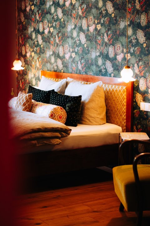 Hotelkamer met bloemenbehang, houten hoofdbord in okergeel damast en gelaagde kussens in zwart, wit en oker, door cestsjoukje