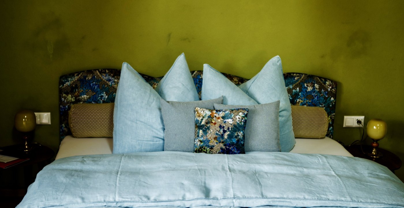 Hotelkamer met gebloemd hoofdbord in stof van Rohleder tegen een olijfgroene wand in Matcha, met lichtblauw linnen, door cestsjoukje
